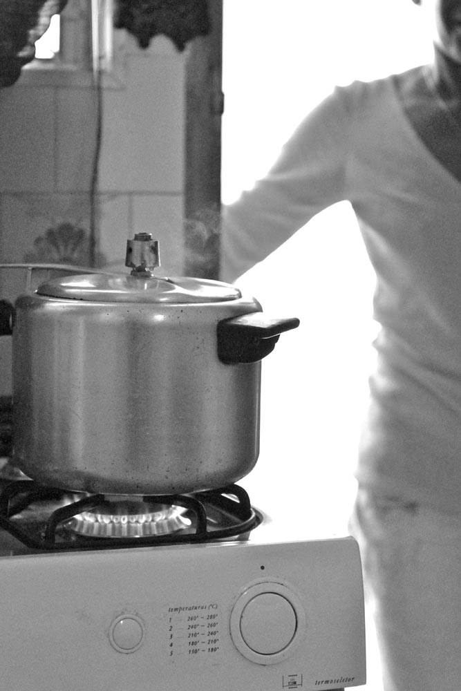 Panela de pressão no fogão e uma figura parcialmente visível ao fundo, na cozinha da casa de Roza.